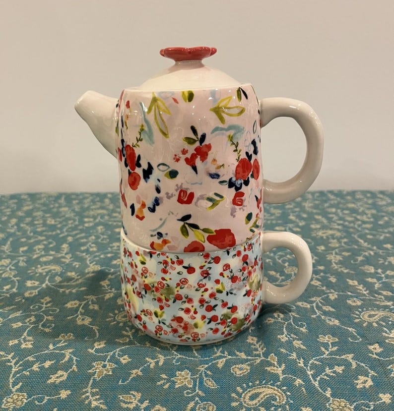 May include: A ceramic teapot and teacup set, stacked together. The teapot is pale pink with a floral pattern of red, blue, and yellow flowers. The teacup has a similar floral design on a light blue background. Both have white handles.