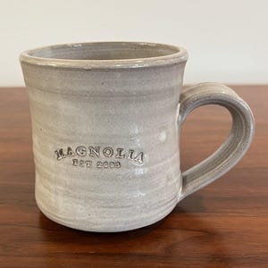 May include: A gray ceramic mug with a curved handle. The mug has the word "MAGNOLIA" and "EST 2003" embossed on the side. The mug is sitting on a wooden surface.