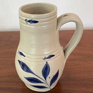 May include: A beige ceramic pitcher with a curved handle and a wide opening. The pitcher is decorated with blue leaf and stem designs. Horizontal ridges encircle the top and middle of the pitcher.
