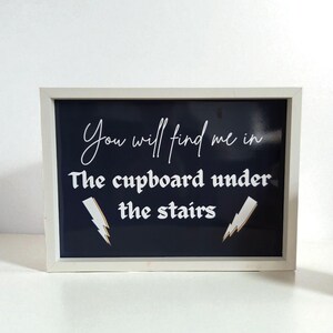 May include: A framed sign with a white border and a dark blue background. The sign features white cursive text that reads "You will find me in" above the phrase "The cupboard under the stairs" in a serif font. Two gold lightning bolt graphics are on the sign.