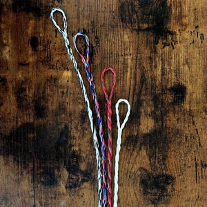 May include: Five colourful twisted cords with looped ends, arranged on a dark wood surface. The cords are white, red, blue, and a combination of these colours. The cords are likely used for crafting or jewellery making.