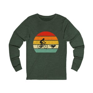 May include: Dark green long-sleeve shirt featuring a retro design of a cyclist and a dog running against a sunset. The graphic includes a cyclist on a bicycle and a dog running to the right. The sunset has horizontal stripes of red, orange, yellow, and green.