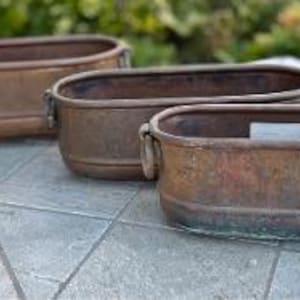 May include: Three oval-shaped copper planters arranged in a row, each featuring a weathered, oxidized patina and two small metal ring handles on the sides. The containers have a smooth, rounded base and a distinct rim, showing signs of age and use.