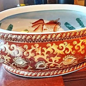 May include: A decorative ceramic bowl with a red and gold floral design. The bowl has gold handles and a white interior with a painted goldfish and aquatic plants. The bowl is likely used for decorative purposes.