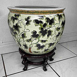 May include: A large, round ceramic planter with a cream-colored base and a dark brown wooden stand. The planter is decorated with a pattern of dark green leaves and vines. The rim has a decorative gold and green border.