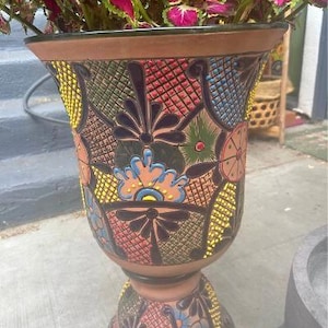 May include: A tall, flared terracotta planter featuring intricate hand-painted geometric and floral patterns in vibrant shades of yellow, blue, green, and red. The surface is divided into sections with black outlines, and a plant with pink and green leaves sits inside.