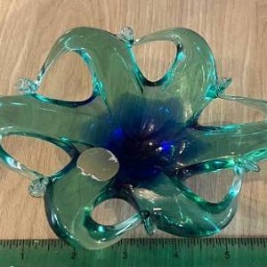 May include: A hand-blown glass bowl featuring a star-shaped design with six elongated, curved arms. The glass transitions from a deep, saturated blue at the center to a translucent teal green at the edges. A small circular sticker is visible on one arm.