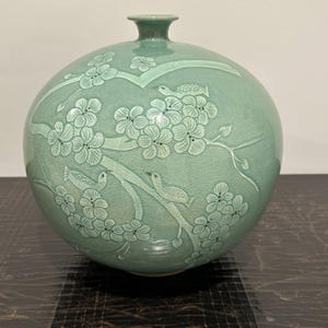 May include: A celadon-colored ceramic vase with a round body and a narrow neck. The vase features an embossed design of flowering branches and birds. The overall aesthetic is elegant and traditional, suitable for home decor.
