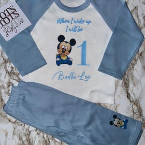 Pode incluir: Um conjunto de pijama para bebé azul claro e branco. A parte de cima tem mangas azuis e um torso branco com um desenho do Mickey Mouse e o texto "When I wake up I will be 1". As calças a condizer são azuis claras com um desenho do Mickey Mouse.