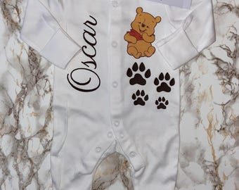 Könnte beinhalten: Weißer Baby-Strampler mit langen Ärmeln, mit Winnie Puuh-Grafik, Pfotenabdrücken und dem Namen "Oscar" in brauner Schrift. Der Strampler hat Druckknöpfe und ein "Tots Babywear"-Etikett.