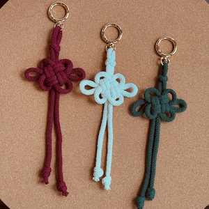 May include: Three handmade keychains in shades of burgundy, light blue, and dark green. Each keychain features a decorative knot design with dangling cords and a gold-colored ring for attachment. The keychains are displayed on a neutral-toned surface.