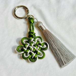 May include: A green, white, and silver keychain with a decorative knot design and a silver tassel. The keychain has a silver clasp for attaching to keys or bags. The knot is a flower-like shape.