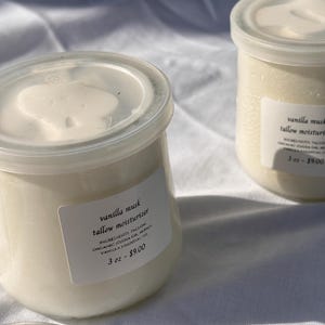 Op de afbeelding: Twee kleine, doorzichtige potjes met witte deksels, gevuld met een romige, crèmekleurige moisturizer. Elk potje heeft een etiket met de tekst "vanilla musk tallow moisturizer". De potjes staan op een wit oppervlak.