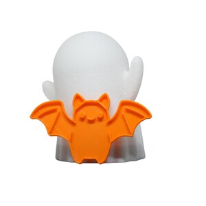 May include: A white ghost figure with an orange bat attached to its front. The bat has a smiling face and outstretched wings. The Halloween-themed decoration is set against a white background.