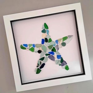 May include: A framed artwork featuring a starfish design. The starfish is composed of various shades of green, blue, and white sea glass pieces. The artwork is set against a white background within a white frame with a black inner border.