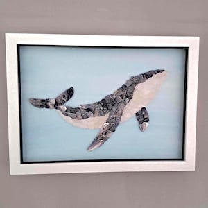 May include: A framed wall art piece featuring a whale mosaic crafted from small grey and white stones. The whale is positioned against a light blue background, encased in a simple white frame with a black inner border, creating a clean display.