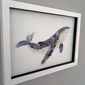 Peut inclure: Une œuvre d'art murale encadrée représentant la silhouette d'une baleine composée de divers éclats de verre de mer et de galets dans des tons gris et blancs. La baleine est placée sur un fond gris clair dans un cadre rectangulaire blanc.