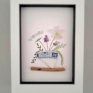 May include: Framed artwork depicting a floral arrangement crafted from sea glass and pottery fragments. The flowers are white, purple, and green, presented in a blue and white patterned vase. The artwork is set against a white backdrop within a white frame.