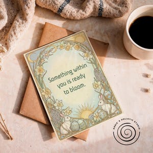 Op de afbeelding: Een wenskaart met de tekst "Something within you is ready to bloom." De kaart heeft een bloemenrand met narcissen, konijnen en paaseieren. De kaart ligt op een bruine envelop, met een kopje koffie op de achtergrond.