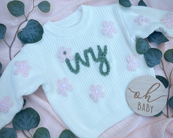 May include: A cream-coloured knit sweater featuring the name Ivy embroidered in dark green yarn across the chest. The sweater is decorated with several small, light pink yarn flowers and includes a round wooden plaque with the text oh BABY.