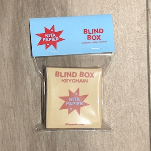 Peut inclure: Un petit porte-clés blind box carré dans un sac en plastique transparent. La boîte est beige avec des graphiques en forme d'éclat d'étoile rouge et le texte "BLIND BOX KEYCHAIN" et "NITA PAPIER". Une carte bleue avec des graphiques et du texte assortis est au-dessus.