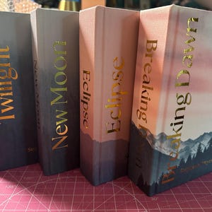 Puede incluir: Cuatro libros de tapa dura con letras doradas. Los títulos son "Twilight", "New Moon", "Eclipse" y "Breaking Dawn". Las portadas de los libros tienen un diseño degradado con una escena de montaña en el último libro.