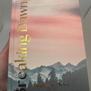 May include: Hardcover book titled "Breaking Dawn" with gold lettering. The cover features a landscape of mountains and trees under a pink and purple sky. The author's name, Stephanie Meyer, is also in gold.
