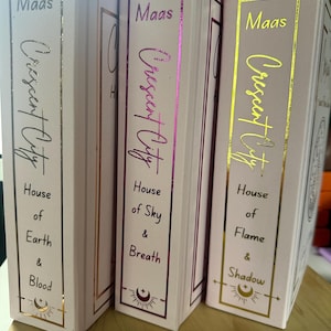 Puede incluir: Tres libros de tapa dura con cubiertas rosas y letras doradas o moradas. Las lomos dicen "Maas" y "Crescent City" con títulos como "House of Earth & Blood". También hay marcos y símbolos dorados decorativos.