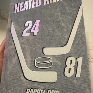 Peut inclure: Couverture de livre avec le titre "Heated Rivalry" en lettres irisées. La couverture présente des crosses de hockey, un palet et les chiffres 24 et 81. Le nom de l'auteur, Rachel Reid, est imprimé en bas.