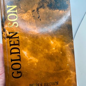 Op de afbeelding: Een hardcover boek getiteld Golden Son van Pierce Brown. De kaft heeft een levendig oranje en zwart nevelontwerp met verspreide witte sterren. De titel is gedrukt in grote, verticale gouden letters aan de linkerkant, auteur in goud.