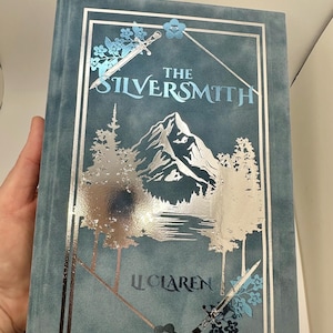 Puede incluir: Un libro de color turquesa titulado "The Silversmith" con detalles en lámina plateada. La portada presenta una escena de montaña, árboles y espadas. El nombre del autor, "LL Claren", está impreso en la parte inferior. El libro tiene una textura aterciopelada.