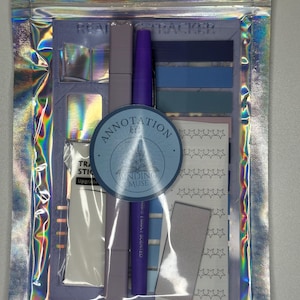 Puede incluir: Un kit de anotación en una bolsa holográfica que contiene un marcador morado, un resaltador morado claro, una hoja de pegatinas de estrellas y varias banderas adhesivas azules. Una etiqueta circular frontal dice Annotation Kit Binding Muse.