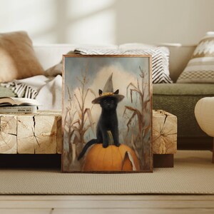 May include: A rectangular framed wall art print featuring a black cat wearing a pointed witch hat with a yellow band, sitting atop a large orange pumpkin in a field of dried corn stalks under a soft, muted sky with light blue and beige tones.