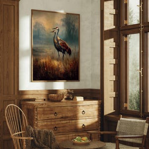 May include: A framed wall art print featuring two cranes standing in a marshy landscape with tall golden grasses, misty trees, and a soft sunset sky. The artwork is displayed on a white wall above a wooden dresser in a room with wood paneling.