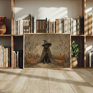 May include: A framed rectangular wall art print featuring a painting of a black dog wearing a pointed witch hat and a cape, sitting in a field of dried corn stalks and orange pumpkins. The artwork has a muted, earthy color palette and soft lighting.