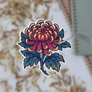 May include: A sticker featuring a stylized chrysanthemum flower. The flower has red, orange, and yellow petals with dark blue-green leaves. The sticker is held by tweezers against a blurred background.