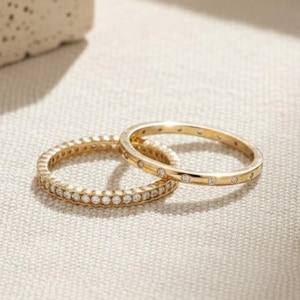 May include: Two gold rings. One ring is fully encrusted with small, round, white gemstones. The other ring has a series of small, round, white gemstones set into the band. The rings are on a neutral-coloured surface.
