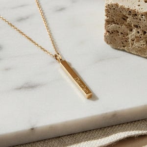 May include: A gold-coloured bar pendant necklace with the name "Damien" engraved on it. The necklace is displayed on a white marble surface, next to a textured, beige stone.