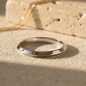 May include: A silver ring with a flat top and angled sides. A gold chain is draped over a beige stone in the background. The ring is set on a neutral-colored fabric.