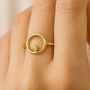 May include: A gold-colored ring featuring a circular design with a small, clear gemstone set in the center. The ring is shown on a person's finger, highlighting its delicate and elegant appearance. The ring's design is simple yet eye-catching.