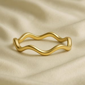 May include: A gold-colored ring with a wavy design. The ring is made of a shiny metal and sits on a cream-colored fabric. The ring's design features a series of smooth, undulating curves, creating a fluid and elegant appearance. The ring is a piece of jewelry.