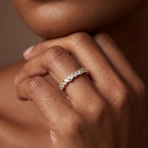 May include: A gold ring with a series of clear, marquise-cut gemstones set in a row. The ring is on a person's finger, with the skin tone appearing warm and natural. The ring's design is elegant and suitable for various occasions.