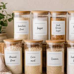 May include: Ten clear glass cylindrical storage canisters with bamboo lids, arranged in two rows. Each canister features a white rectangular label with black text identifying contents: Semolina flour, Sugar, Dark Brown Sugar, Powder Sugar, Mexican Insta Corn flour, Almond flour, Corn meal, Quinoa, Insta Corn flour, and Light Brown Sugar.