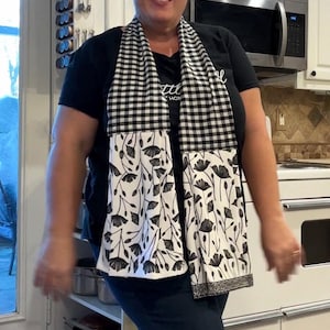 May include: A black and white kitchen towel scarf with a gingham top and a floral pattern on the bottom. The scarf is draped around the neck. The background shows a kitchen with white cabinets and appliances.