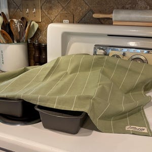May include: Two dark gray loaf pans are covered with a light green striped kitchen towel on a white stove. A rolling pin rests above the stove. Kitchen utensils are in a white ceramic container.