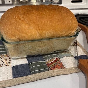 May include: A golden-brown loaf of bread in a clear glass baking dish rests on a patchwork patterned hot pad. A wooden bread knife is positioned to the right. The scene is set on a white stovetop.