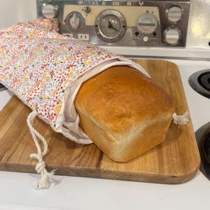 May include: A loaf of golden-brown bread rests on a wooden cutting board, partially enclosed in a floral fabric bread bag. The bag features a white interior and a drawstring closure. The background includes a vintage stove.