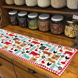 May include: A rectangular farmhouse-style table runner with a white background and a repeating pattern of farm animals, windmills, and other farm-related images in red, blue, and brown. The runner is trimmed with a red and white checkered border.