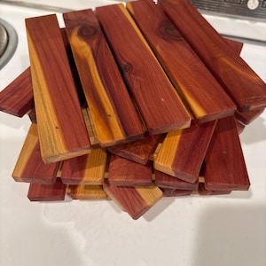 May include: A stack of rectangular cedar wood pieces, displaying a mix of reddish-brown and light yellow-brown hues. The wood grain is visible, and some pieces have knots. The pieces are arranged in a slightly haphazard pile.