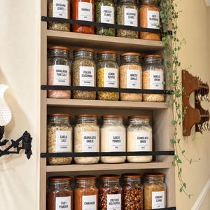 May include: A tall, light-colored wooden spice rack with six shelves holding glass jars with wooden lids and white labels. The jars contain various spices, herbs, and seasonings. A trailing green plant hangs down the right side of the rack.
